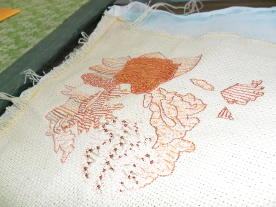 Floral embroidery on pale cloth with reddish outlines and a partly filled orange flower centre